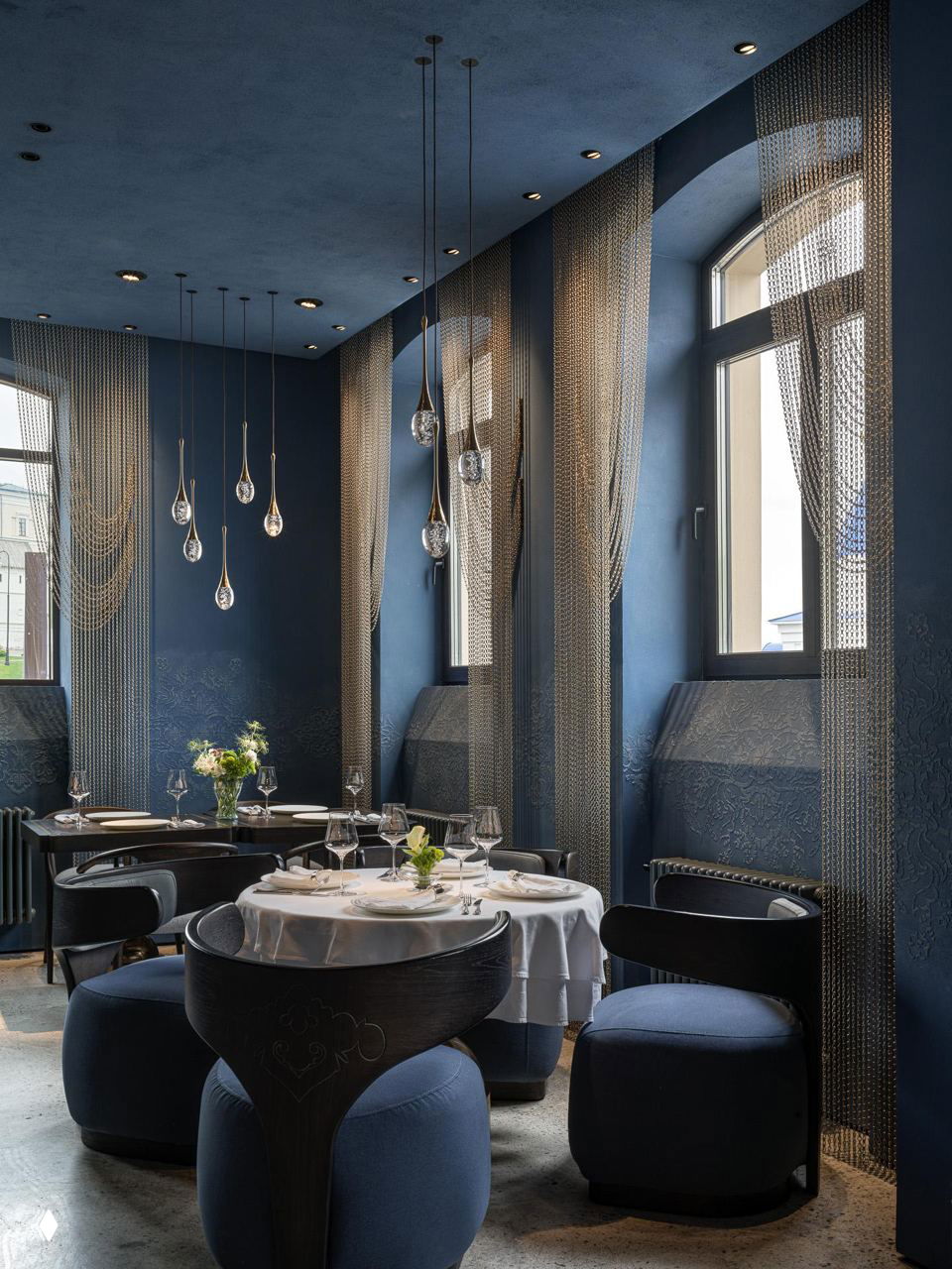 Уголок ресторана с круглыми столами, золотыми подвесными светильниками и высокими арочными окнами на фоне насыщенных синих стен с орнаментом.