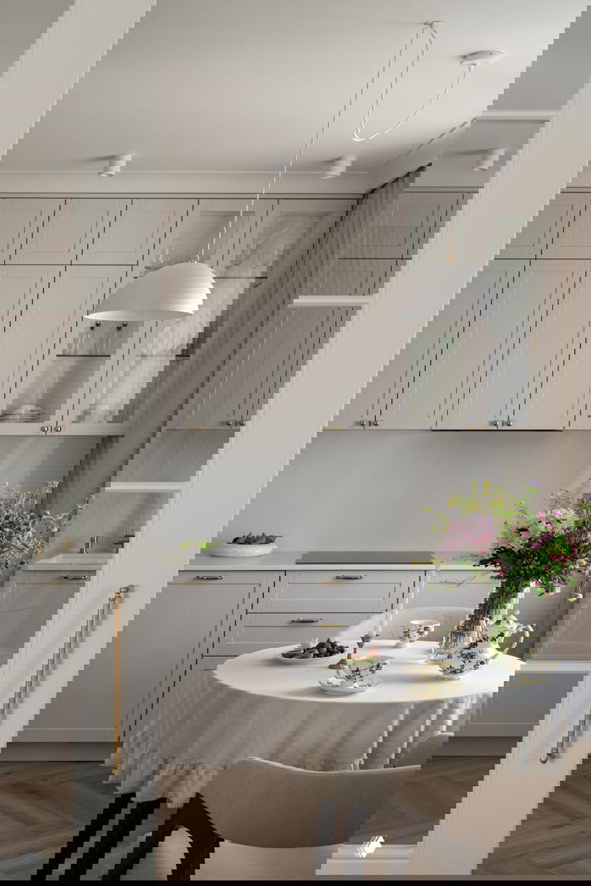 Минималистичная кухня-прихожая в тёплых бежевых тонах, фасады в неоклассическом стиле, стол у окна и подвесной светильник — сочетание сканди и неоклассики.