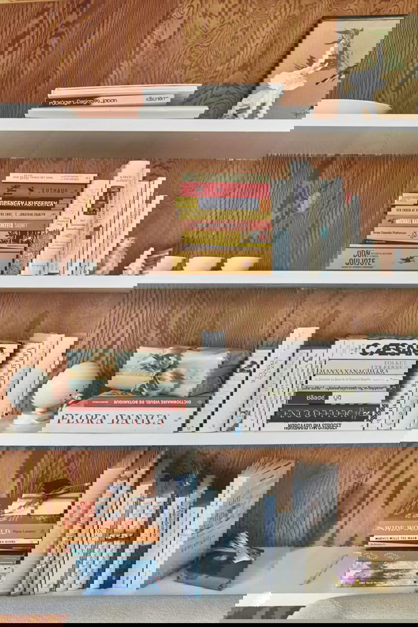 Полка с книгами и декоративными предметами в доме художницы: аккуратно уложенные тома, подарочная упаковка и декоративные фигурки в уютном уголке интерьера