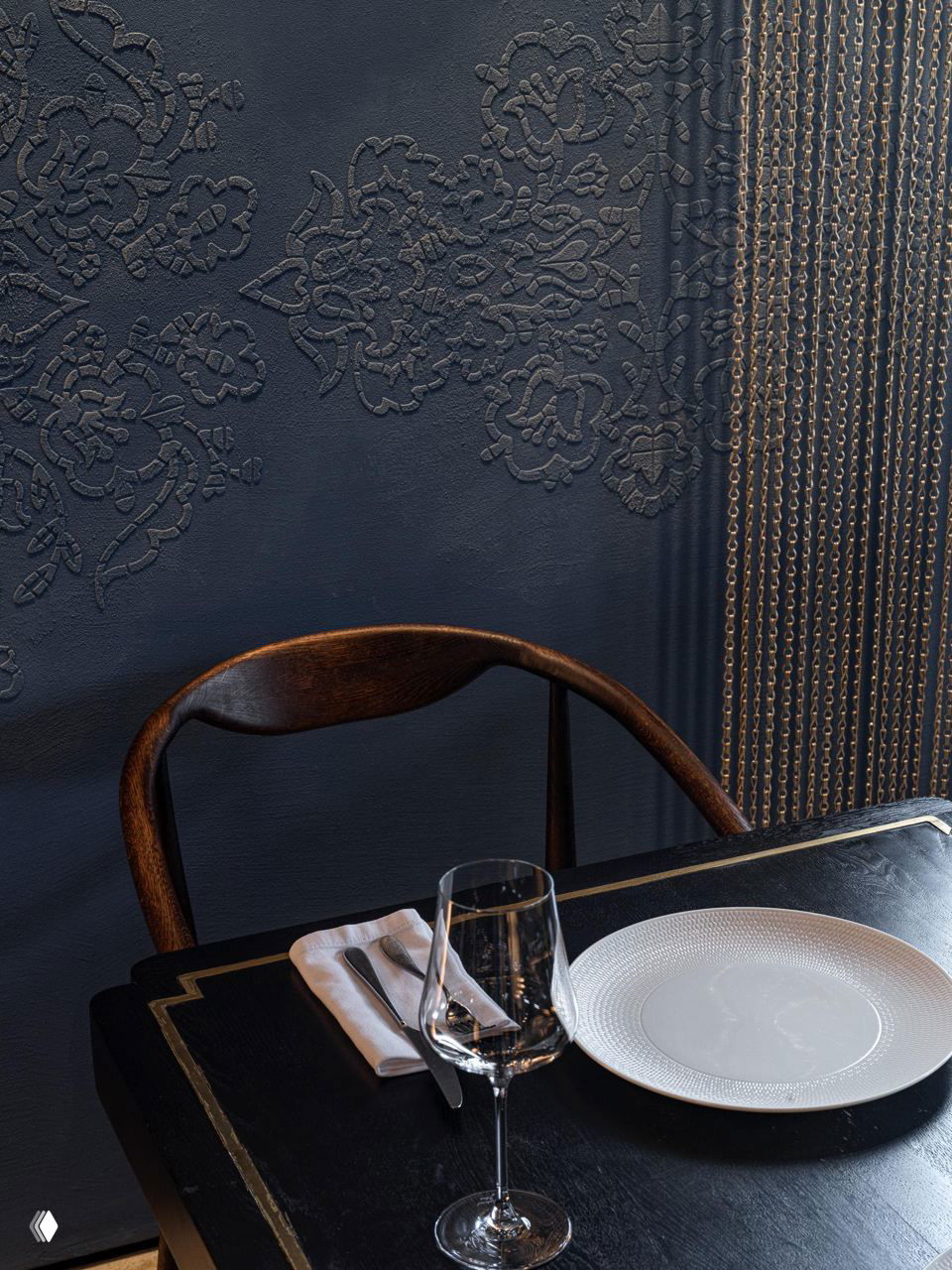 Крупный план текстурной стены с выдавленным татарским орнаментом рядом со столом: тёмно‑синяя фактура, золотые декоративные цепи и часть стула в кадре.