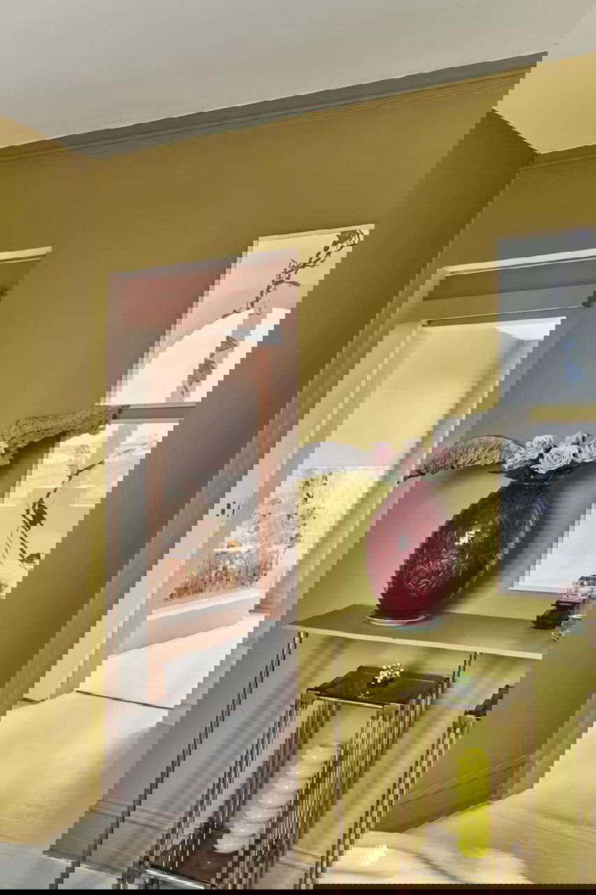 Прихожая с оливково-зелеными стенами, большая красная ваза с цветами и листьями у дверного проема; сквозь дверь видна пастельная зона столовой и розовые акценты