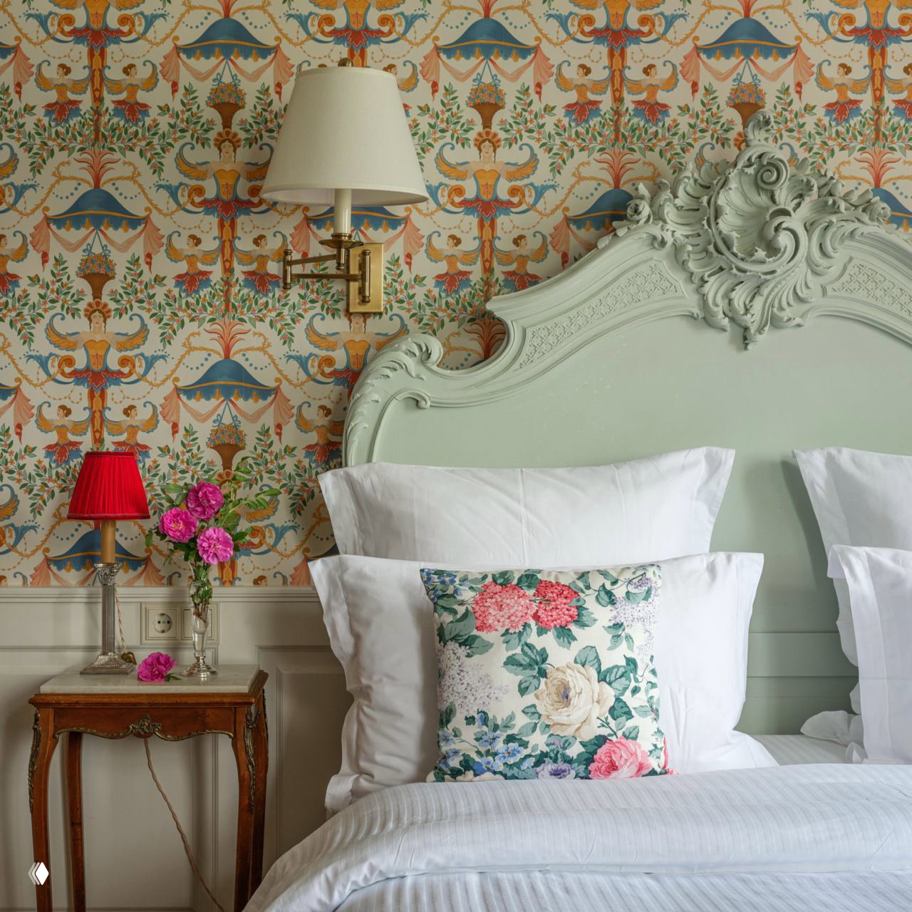 Спальня бутик-отеля с пастельным изголовьем и резными деталями, цветочные обои, белое постельное бельё и декоративная подушка с принтом.