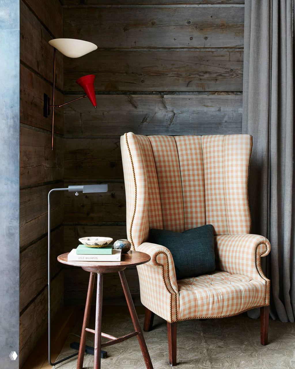 Уютное кресло в клетчатой обивке у деревянной стены, маленький столик с лампой и стопка книг — камерный уголок для чтения.