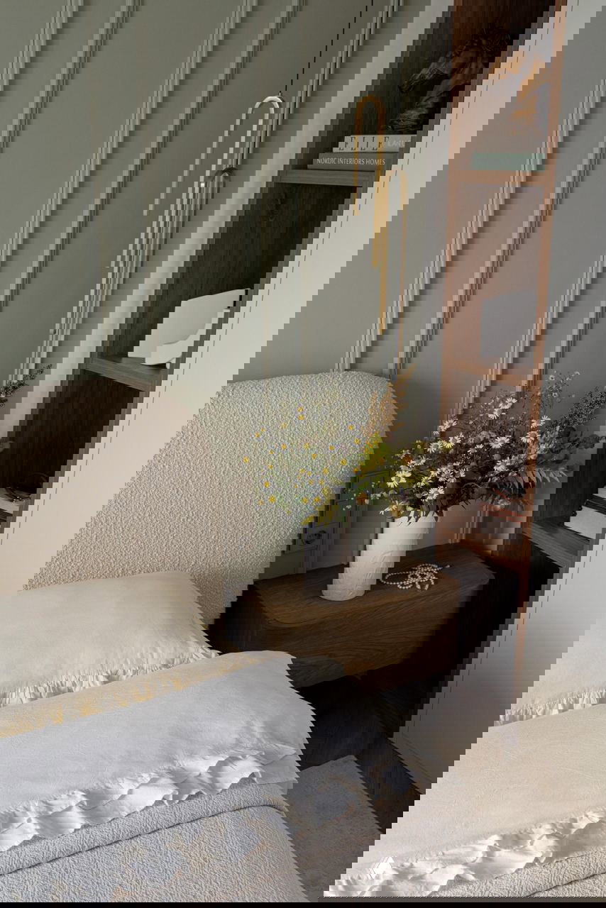 Уютный фрагмент спальни: изголовье в мягкой текстуре, деревянная тумбочка с вазой полевых цветов и вертикальные декоративные панели в спокойном цвете.