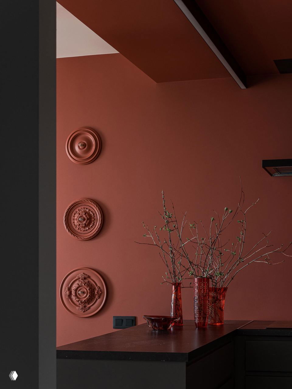 Терракотовая стена с лепными декоративными розетками и красными стеклянными вазами на столешнице, тёмные кухонные фасады создают насыщенный фон.