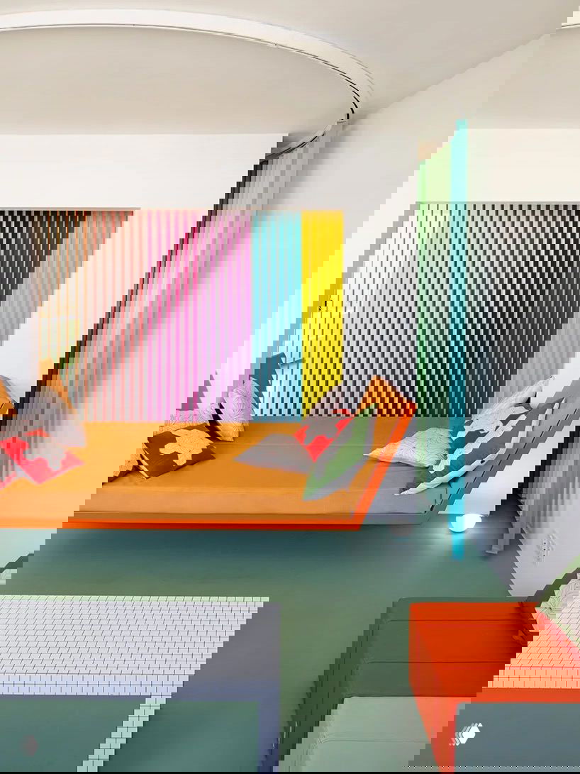 Яркая ниша с вертикальными цветными ламелями и оранжевым диваном‑кроватью — смелое использование цвета и геометрии в интерьере в духе 1980-х.