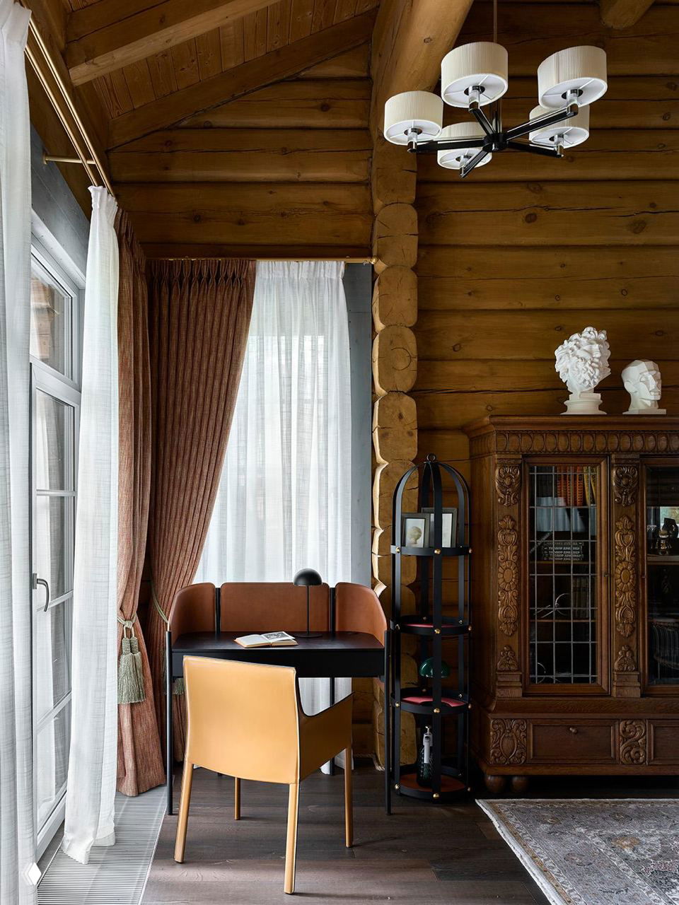 Уютный уголок с рабочим столом, кожаным креслом и резным шкафом на фоне брёвен и высоких окон; проект STone в Подмосковье.