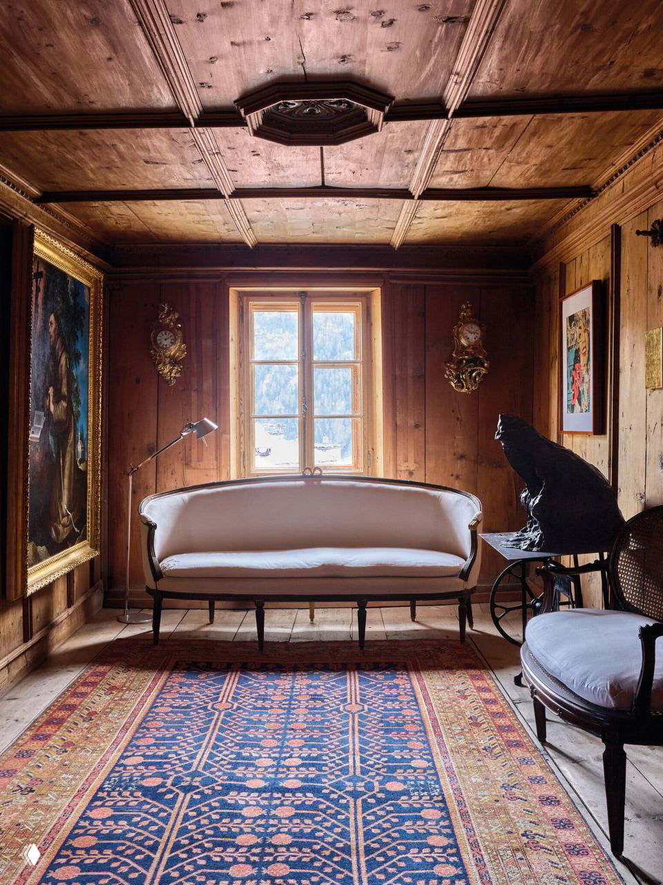 Тёплая комната с панелями из сосны и антикварным диваном в доме Джана Энцо Спероне; деревянный потолок, ковер и окно с видом на швейцарские Альпы создают уют.