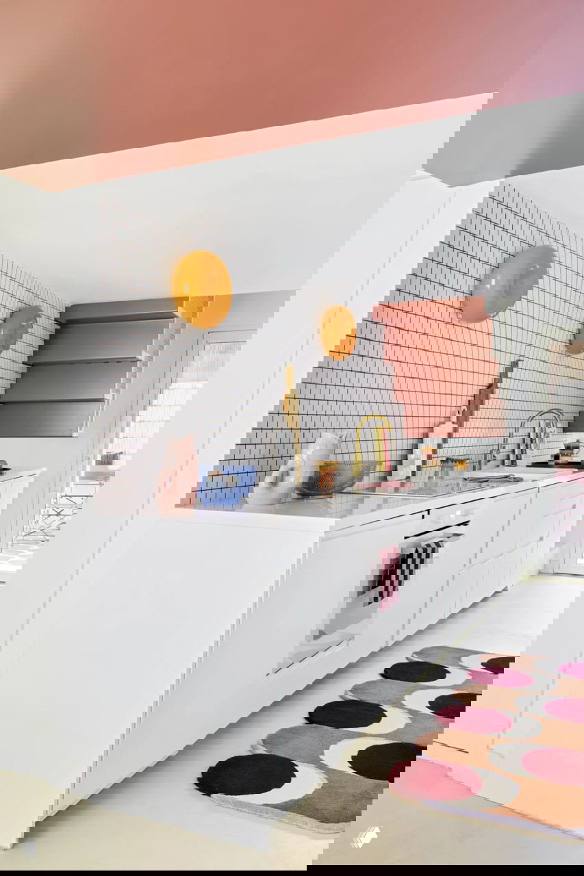 Белая кухня с розовым потолком и настенными декоративными тарелками; розовые ступени и золотистый смеситель формируют поп-арт акценты в интерьере.