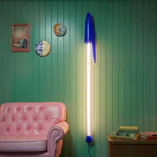 Интерьерная сцена с вертикальным неоновым светильником в синих акцентах, рядом розовый диван и декоративные тарелки на зеленой панели стены.