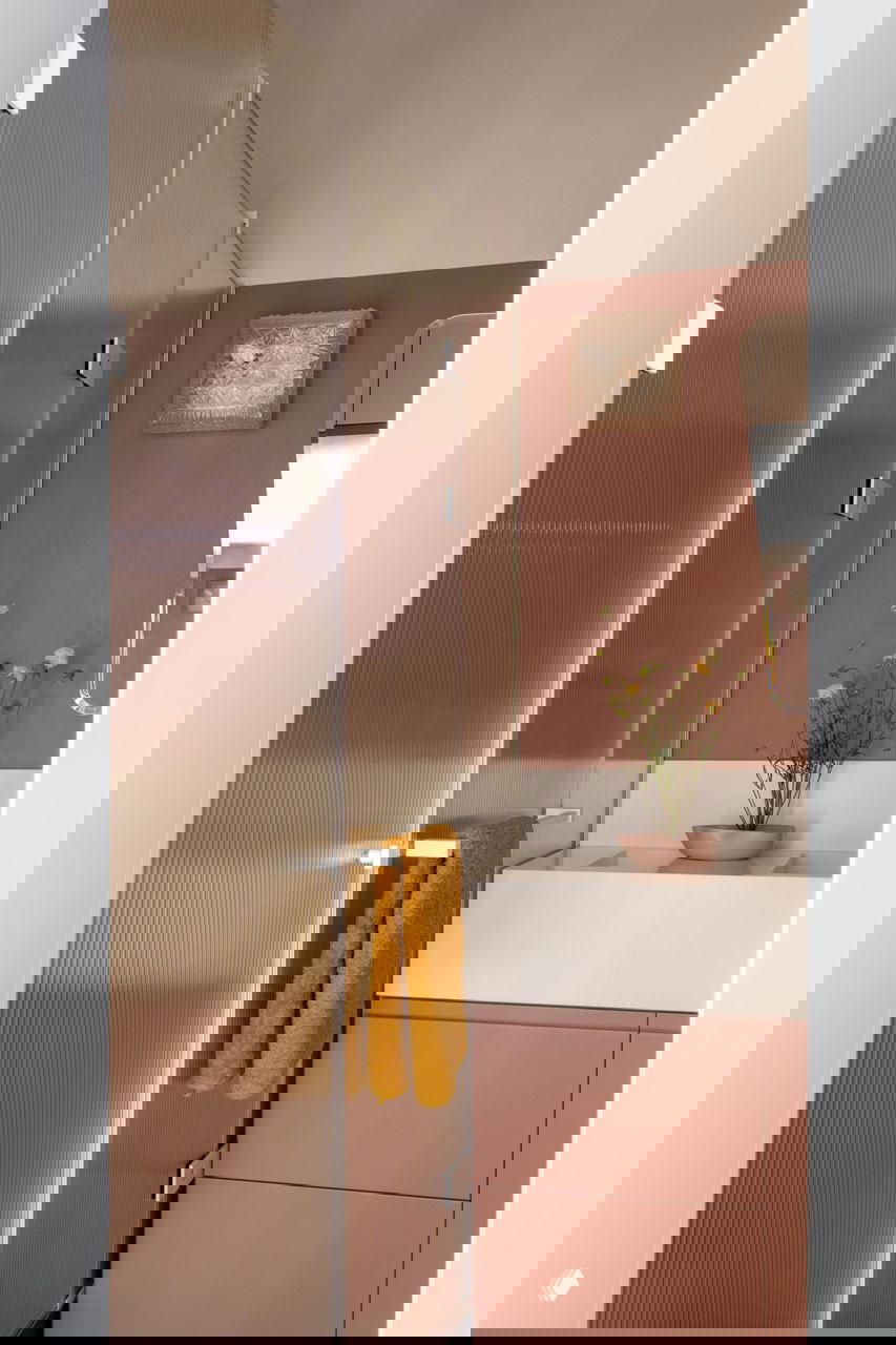 Узкая ванная комната с рифлёной прозрачной перегородкой душа, розово-терракотовые стены и белая прямоугольная раковина с тумбой.