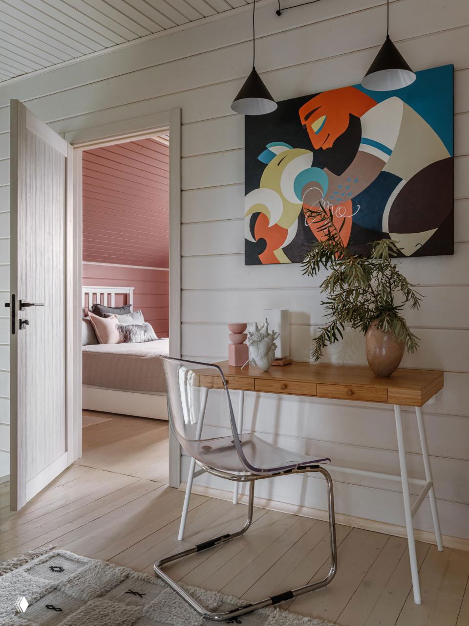 Узкий рабочий стол у стены, прозрачный стул и яркая картина над ним; вид на спальню с розовой отделкой и спокойной палитрой текстиля.