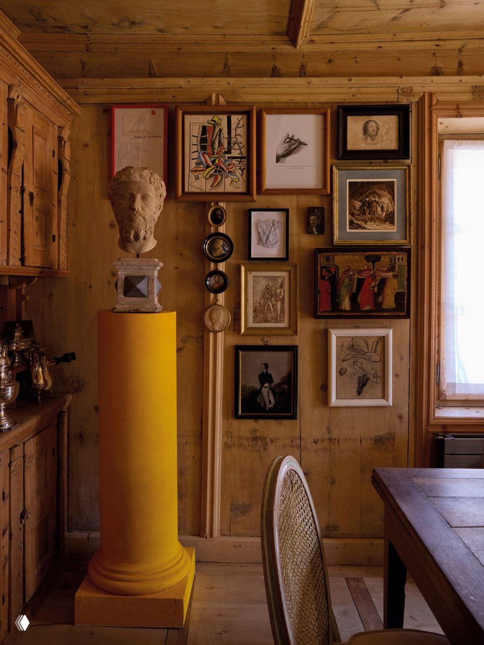 Маленькая рабочая зона с ярко-жёлтым колонновидным пьедесталом, классическим бюстом и множеством рам на деревянной стене — плотная экспозиция мелких произведений.