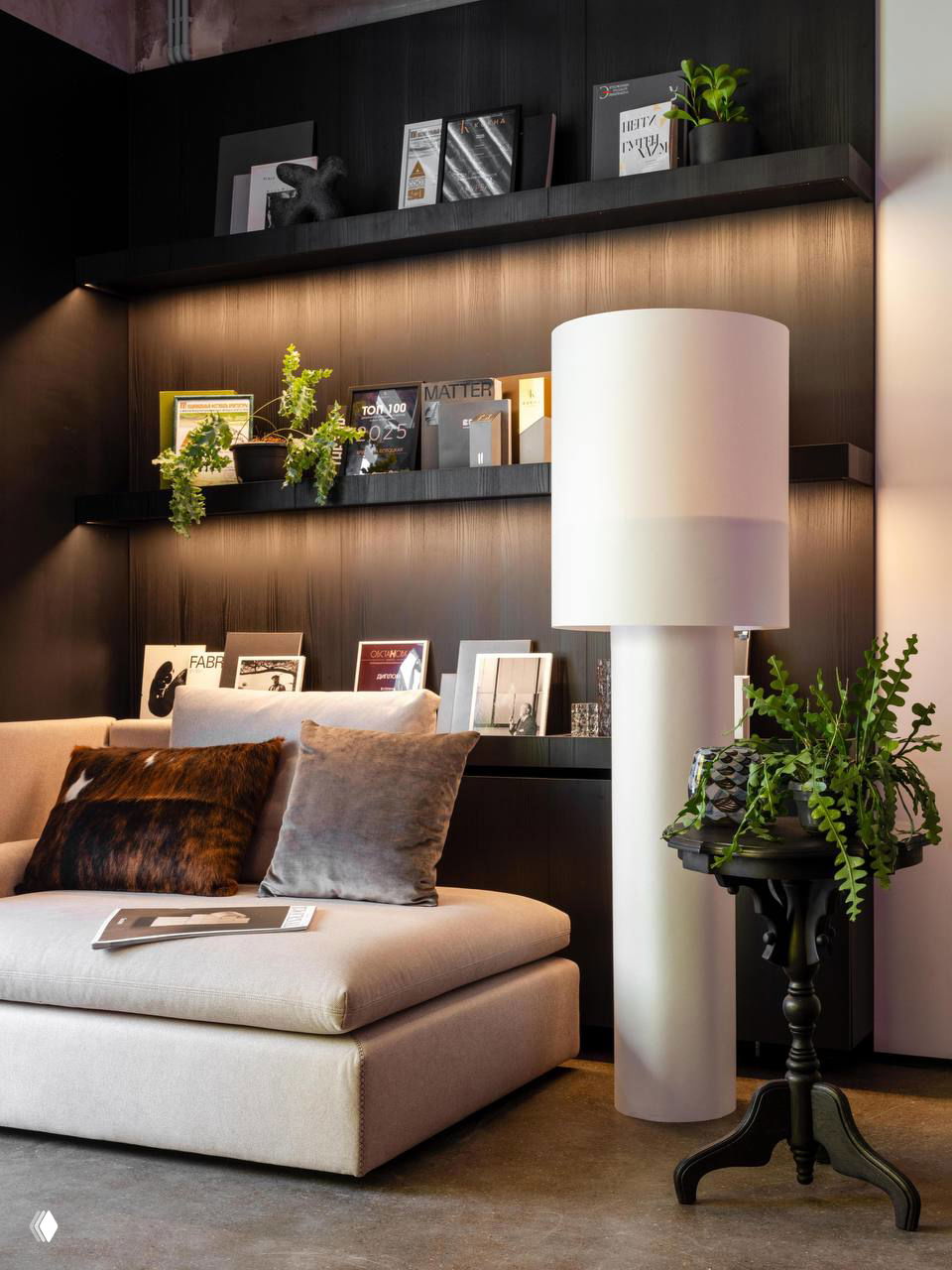 Уютная зона отдыха в офисе: низкий бежевый диван, декоративные подушки, тёмные встроенные полки и аккуратные аксессуары.
