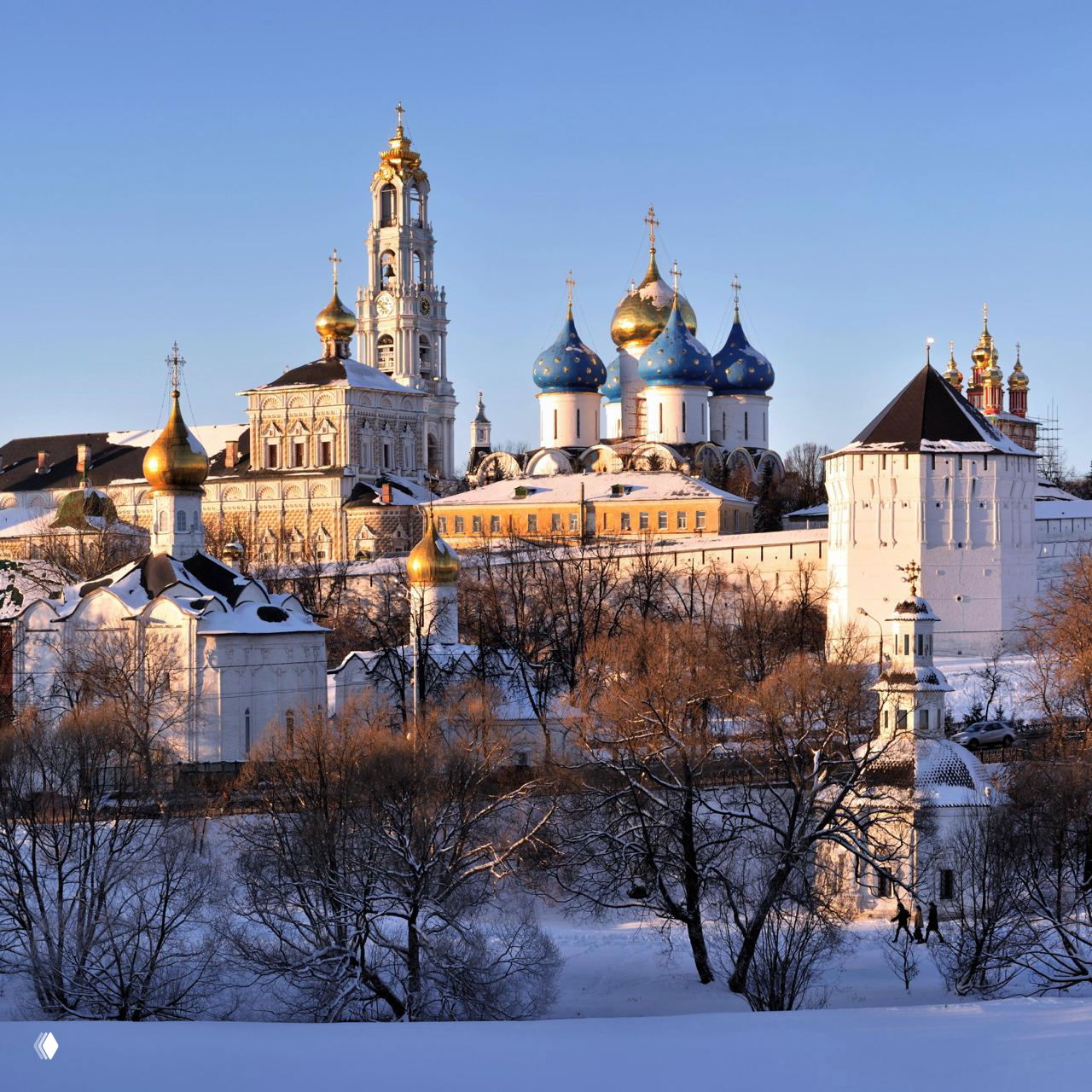 Вид Троице-Сергиевой лавры в зимнем освещении: золотые и синие купола, крепостные стены и заснеженные деревья на переднем плане.