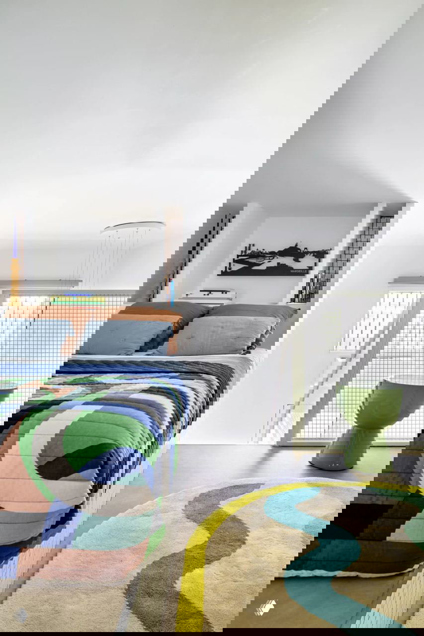 Спальное место на площадке мезонина: разноцветный текстиль, низкий парапет из сетки и зеленый табурет создают уютную антресольную зону.