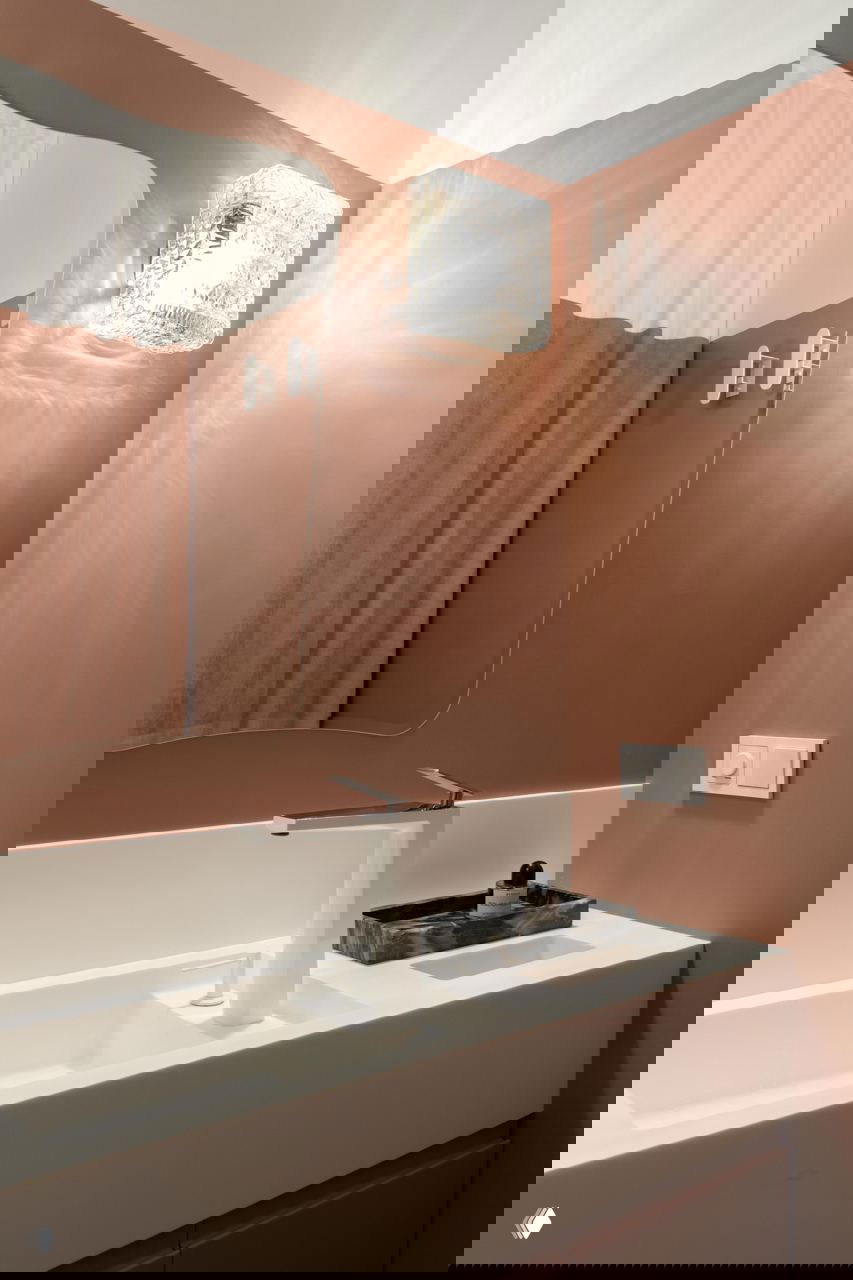 Фрагмент ванной: белая интегрированная раковина, зеркальная панель и настенный стеклянный светильник, отражающийся на розовой стене.