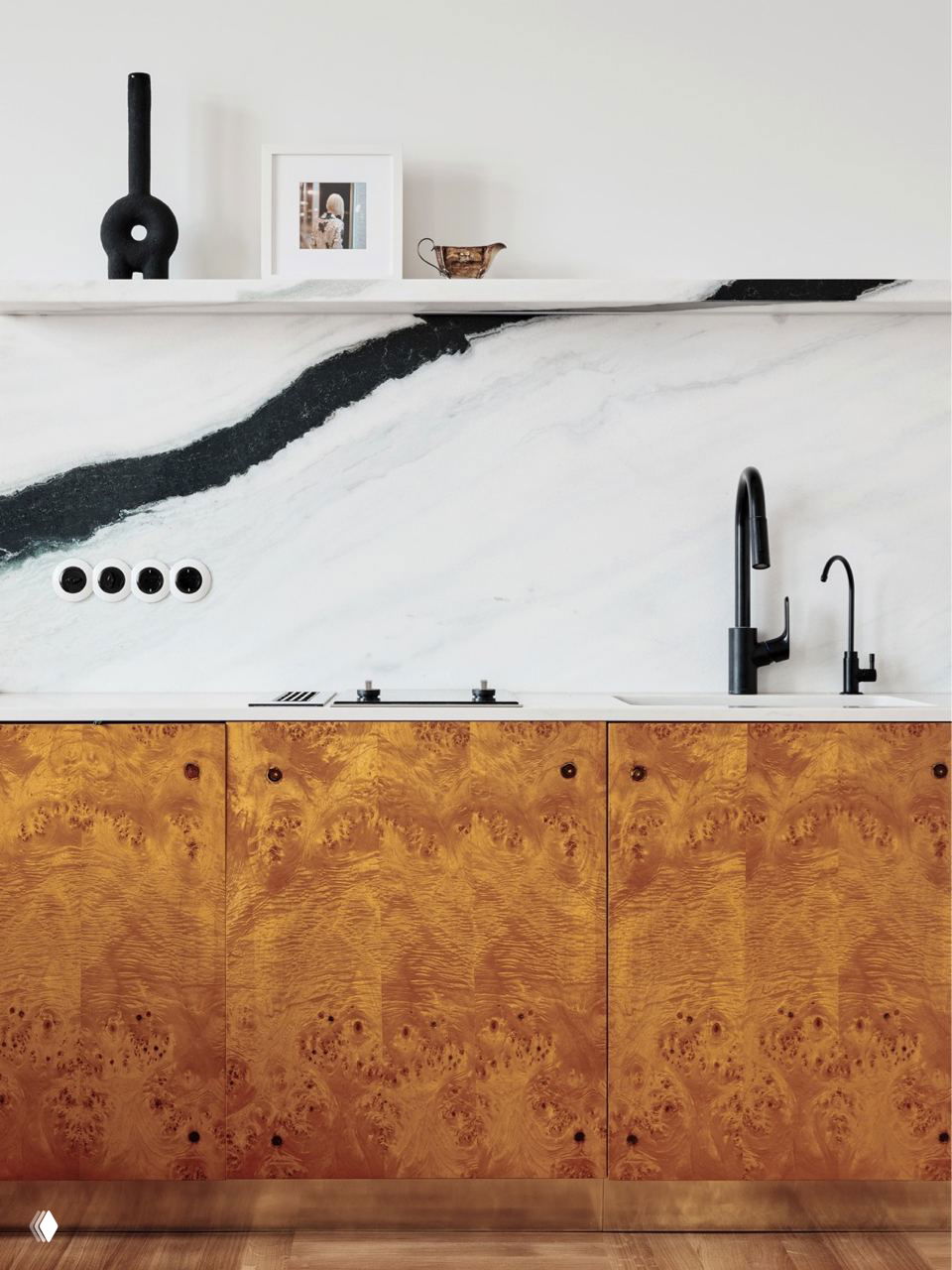 Минималистичная кухонная композиция с белой столешницей, ярко выраженным декоративным рисунком на мраморной стене и нижними фасадами из древесины, без верхних шкафов.