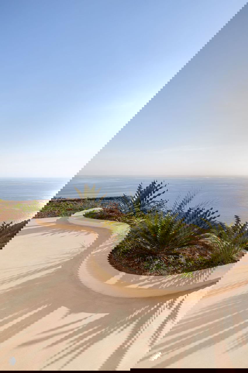 Пейзажный вид с участка дома: изогнутый краевой цветник, сухие растения и открытый горизонт моря на фоне спокойного неба.