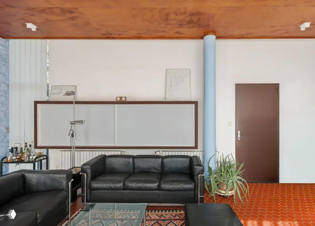 Зона отдыха внутри виллы Ле Корбюзье: черные кожаные диваны, высокий деревянный потолок, терракотовый шестигранный пол и декоративные предметы.