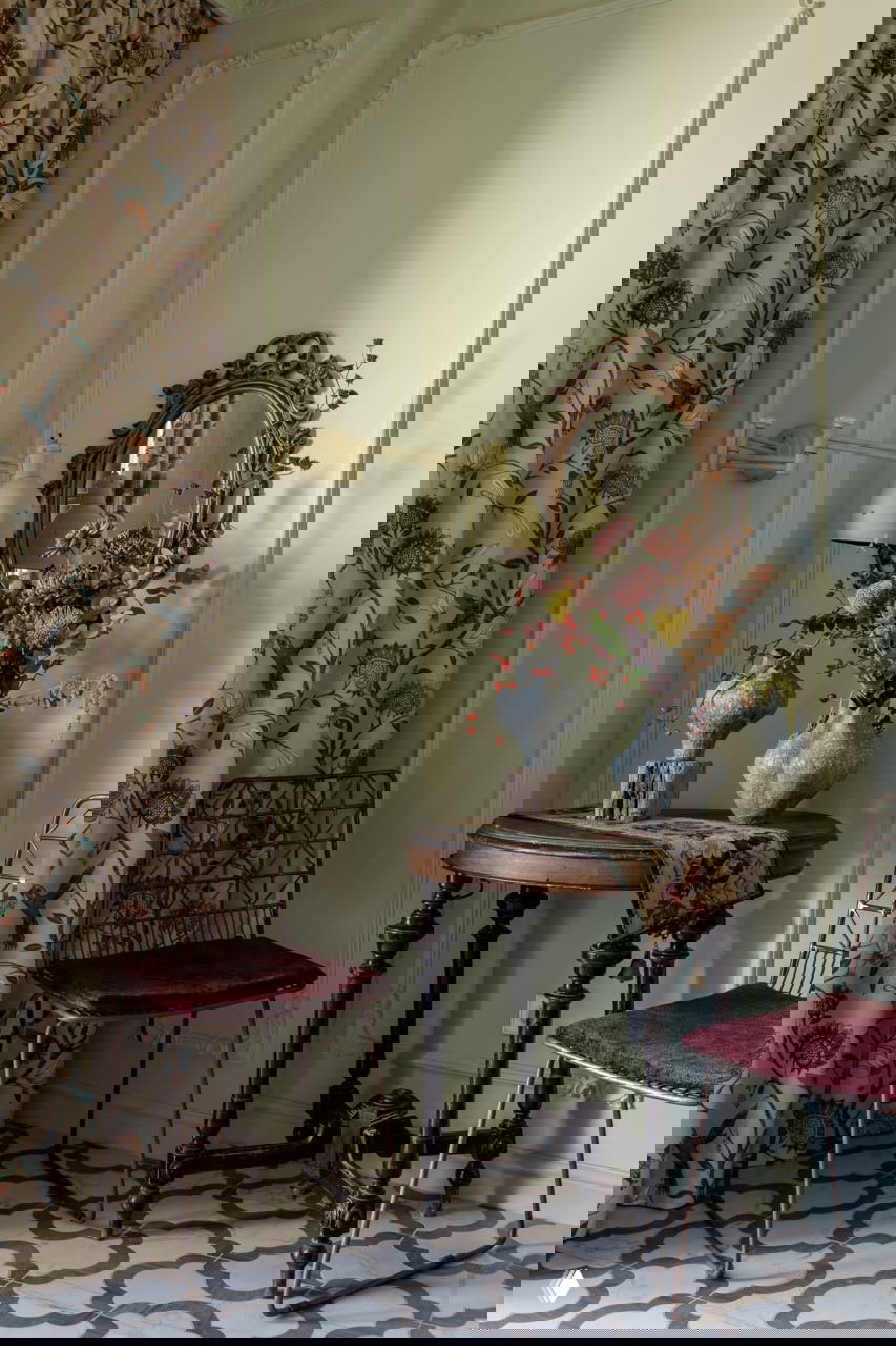 Невысокий круглый стол с вазами и зеркало в резной раме на фоне мягкой зелёной панели и цветочных штор в винтажном стиле.