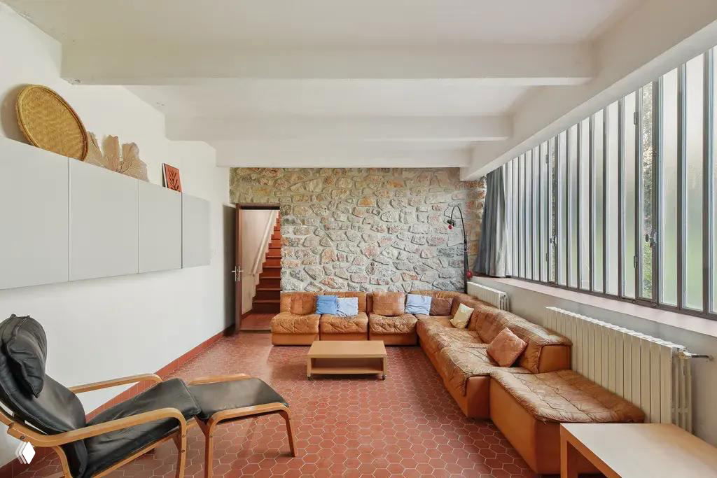 Большая комната с каменной стеной и угловым коричневым диваном в вилле Ле Корбюзье; длинные стальные окна и терракотовый пол создают характерный образ.