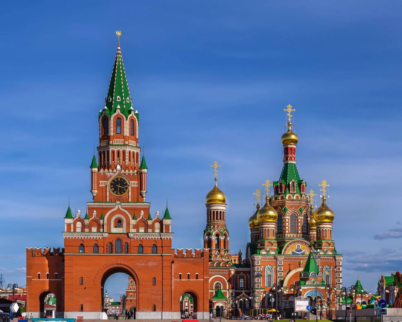 Крупный план уменьшённой копии Спасской башни и куполов собора в том же ансамбле на набережной: красная кирпичная кладка, золотые купола и зелёные шатровые крыши на фоне неба.
