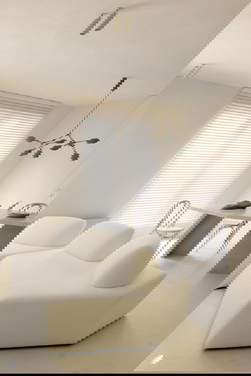 Гостиная с мягким ассиметричным диваном кремового цвета на фоне монохромной стены и вид на обеденную зону с декоративными предметами.