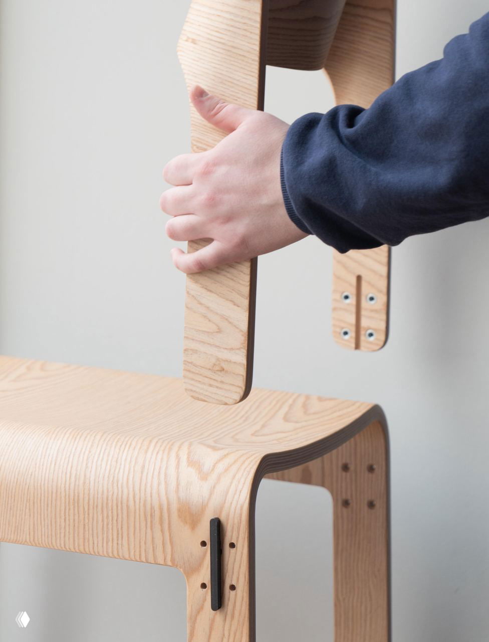 Рука демонстрирует съёмный элемент стула: видно слои гнутой фанеры, отверстия под крепеж и вариант разборной конструкции для замены деталей.