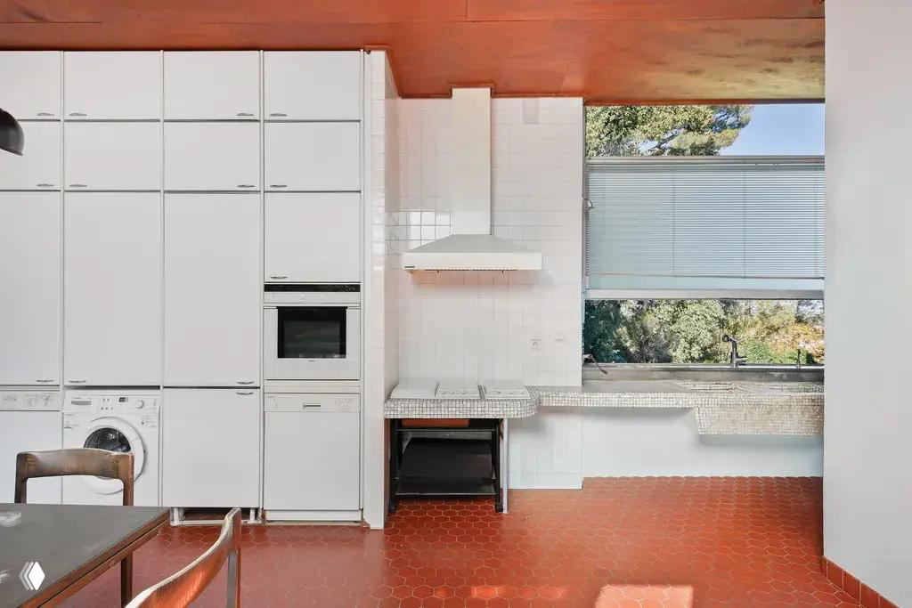 Кухня виллы Ле Корбюзье с высокими белыми шкафами, встроенной техникой и мозаичным рабочим столом; вид на сад через широкое окно обеспечивает много света.