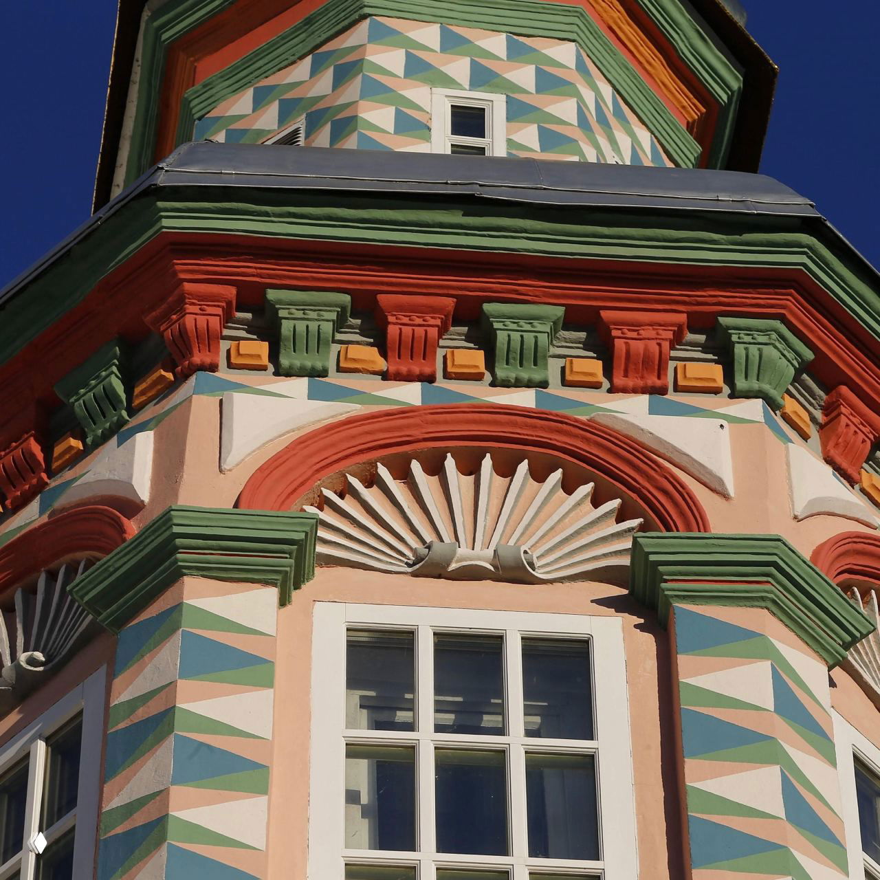 Фрагмент исторического здания с расписным геометрическим фасадом, яркими декоративными элементами и частичным видом окна на фоне чистого неба.