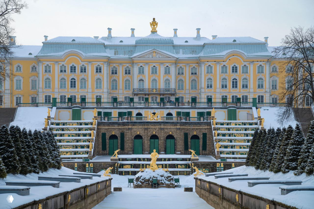 Зимний вид на парадный фасад дворца с золотыми каскадами и заснеженные ступени, архитектура и скульптуры на переднем плане.