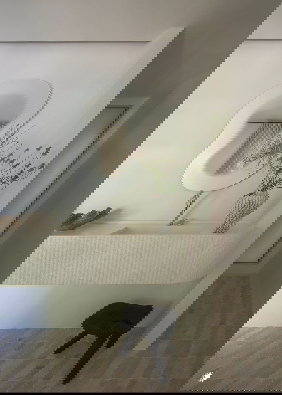 Минималистичная раковина и волнообразное зеркало в прихожей с текстурной вазой и невысоким черным табуретом, спокойная бежевая палитра.