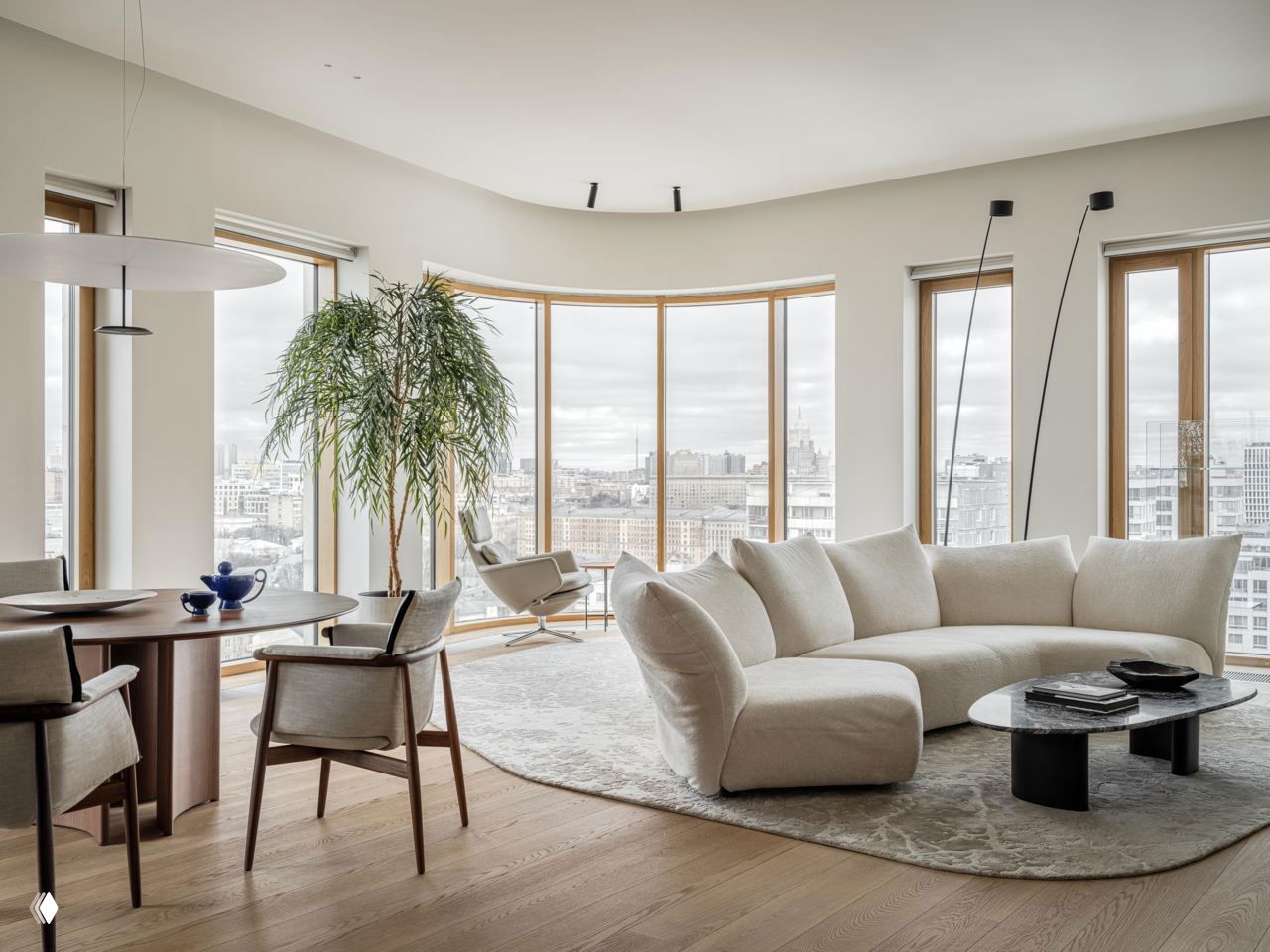 Округлая панорамная стена в гостиной-столовой с деревянными рамами и видом на городской пейзаж; обеденный стол, диван и мягкие оттенки в оформлении.