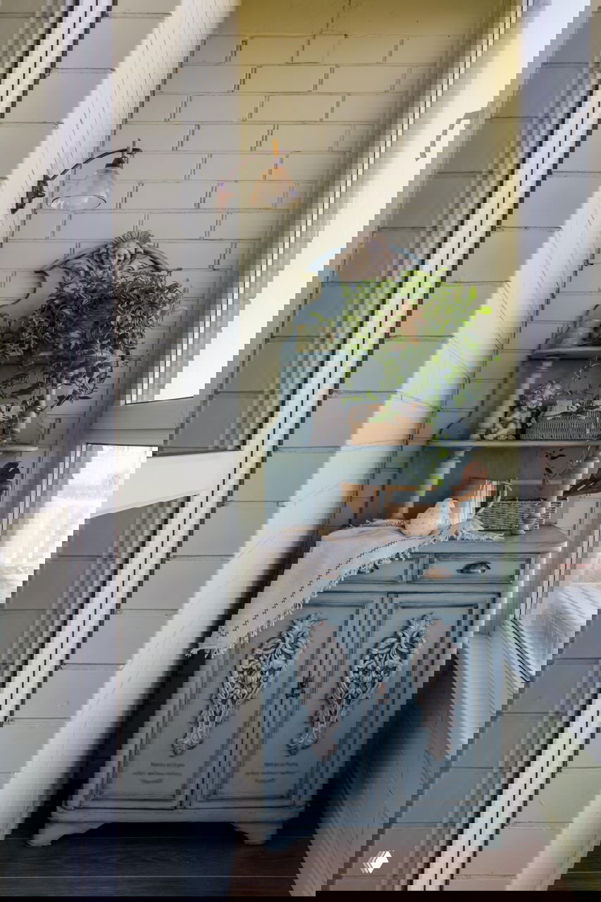 Узкий балкон с голубовато-патинированным шкафчиком в стиле прованс, плетёными корзинами и живыми растениями у окна в солнечном освещении.