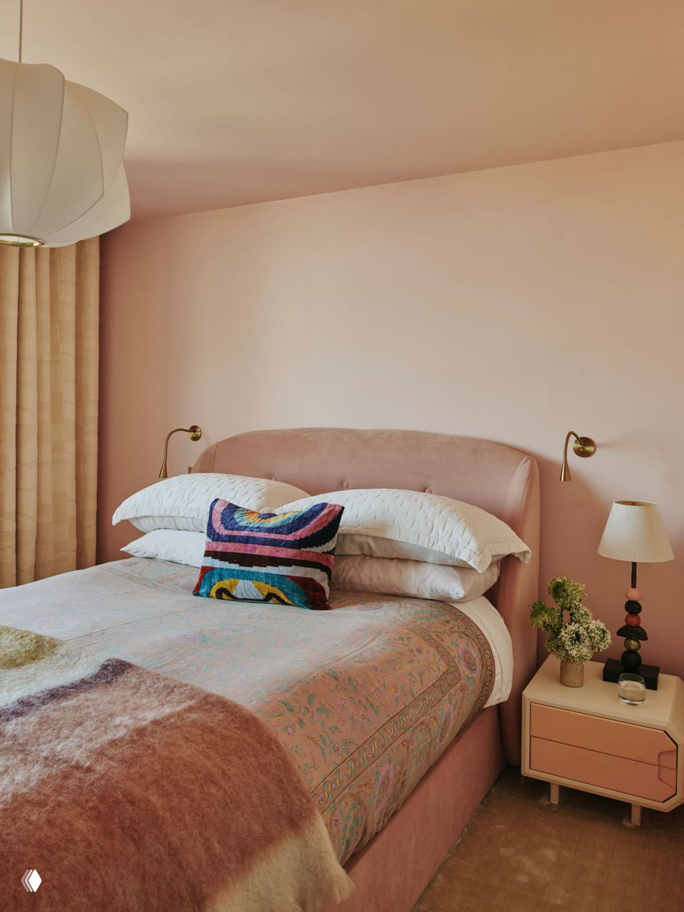 Спальня в пастельных розовых и персиковых тонах: мягкое изголовье, многослойная постель и декоративная подушка с цветным рисунком для баланса ярких акцентов.