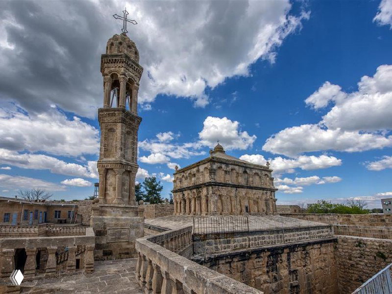 Каменная колокольня и церковное здание в Анытлы на фоне выразительного неба: архитектурные детали и крест на шпиле хорошо видны.