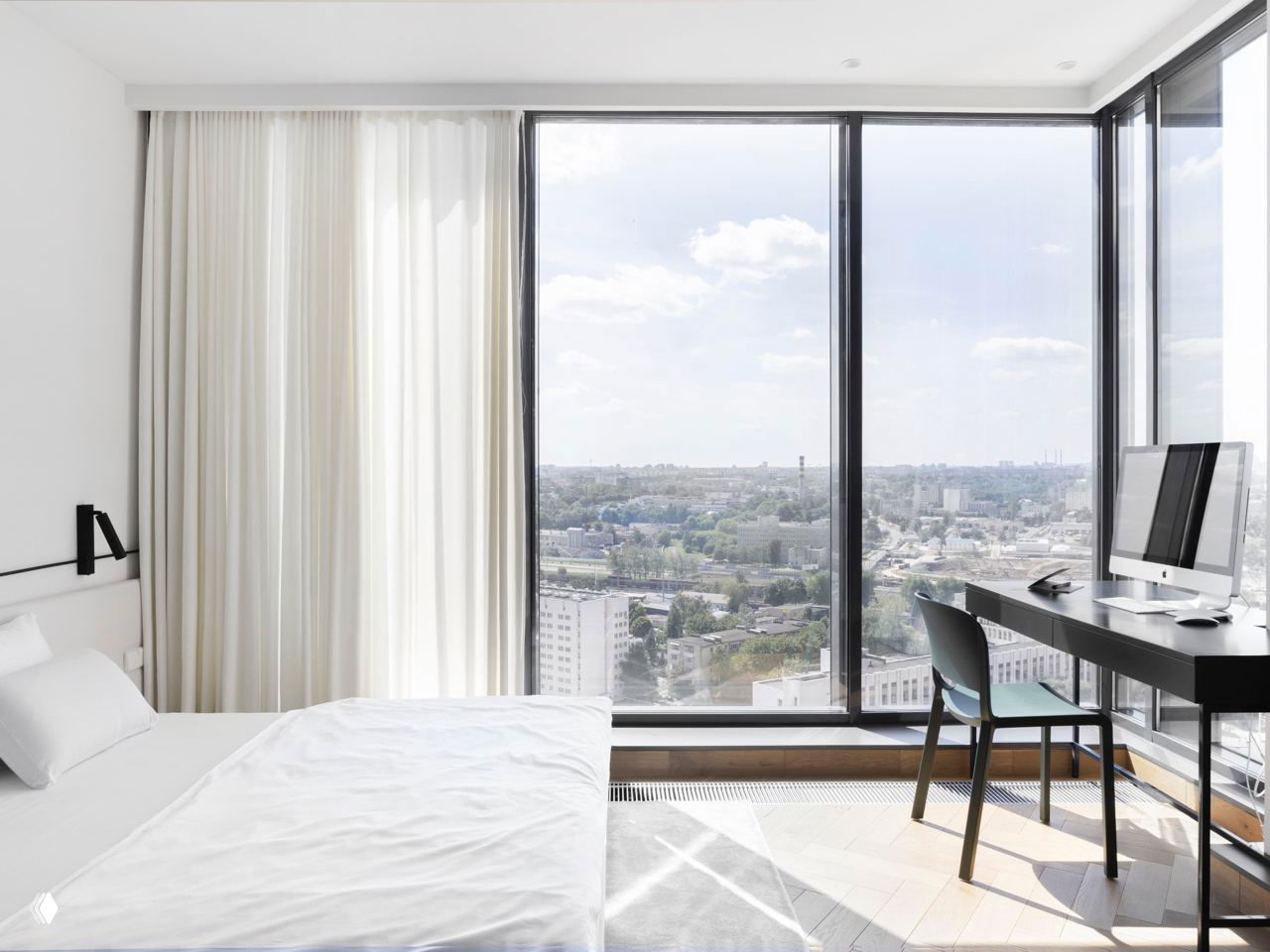 Спальня с панорамным оконным проёмом в пол и рабочим столом у окна; минималистичный интерьер и вид на городские крыши и небо через стекло.