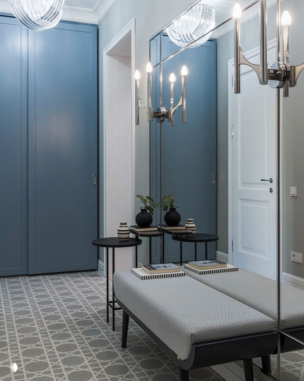 Элегантная прихожая с голубыми встроенными шкафами, зеркальной панелью вдоль стены, пуфом и графичным узором на напольной плитке — сдержанная палитра и аккуратные формы.