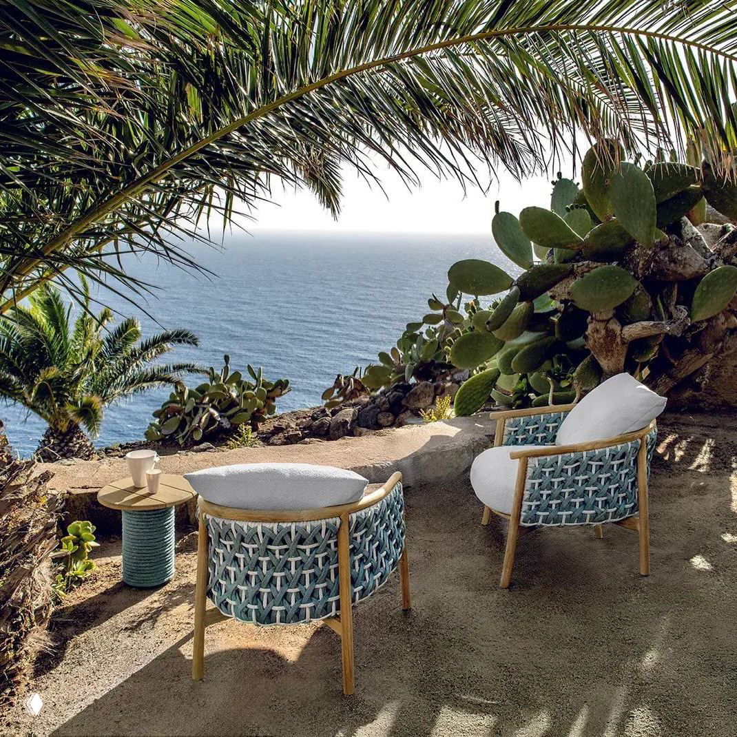 Плотные плетёные кресла с подушками на деревянном каркасе у обрыва с видом на море, пальмы и кактусы в солнечном освещении.