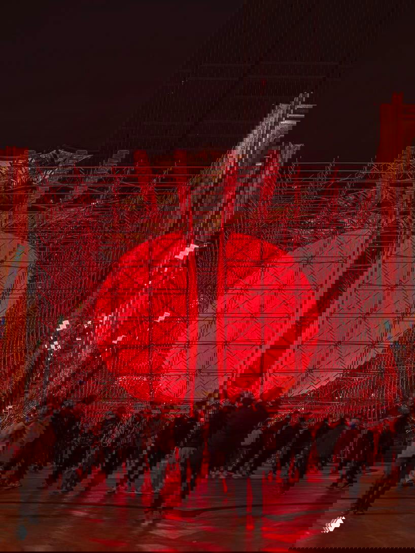 Люди перед большой разделённой красной сферой-инсталляцией: каркас из лесов, яркий свет и толпа на пешеходной зоне вечером.