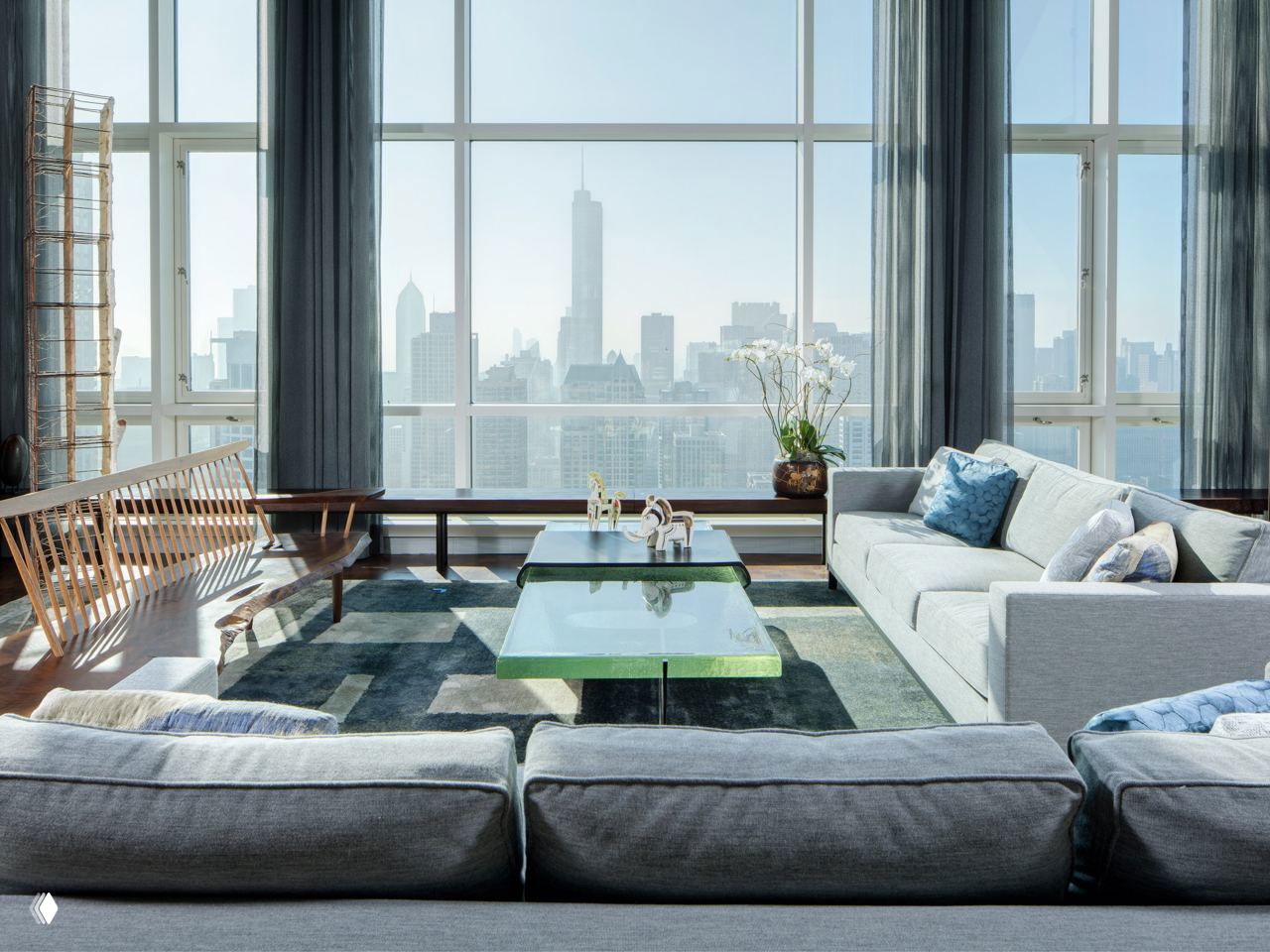 Просторная гостиная с высокими панорамными окнами и видом на силуэт города; серая мягкая мебель и низкий стеклянный стол на декоративном ковре.