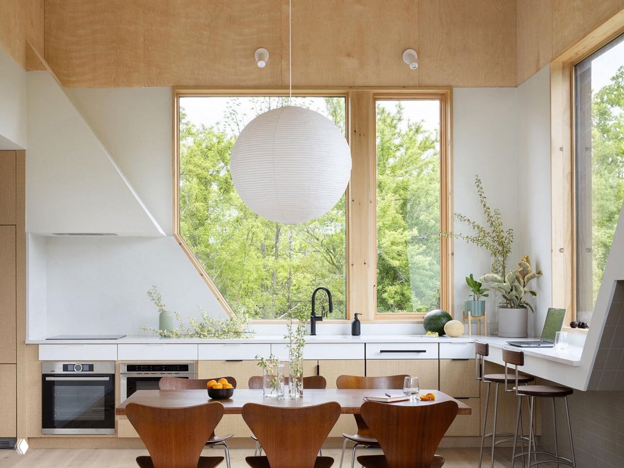 Интерьер кухни с подвесным бумажным светильником над столовой зоной, рабочая стена у окна и светлые деревянные панели в отделке.