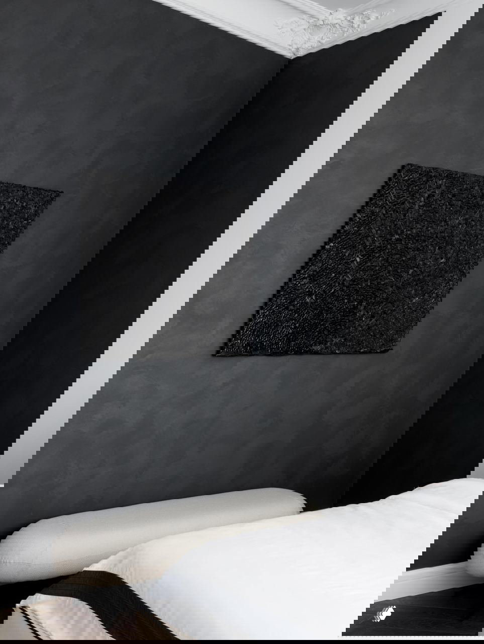 Деталь спальни: фактурная чёрная панель с объёмной картиной, бежевый низкая скамья у кровати и текстиль в спокойной натуральной палитре интерьера