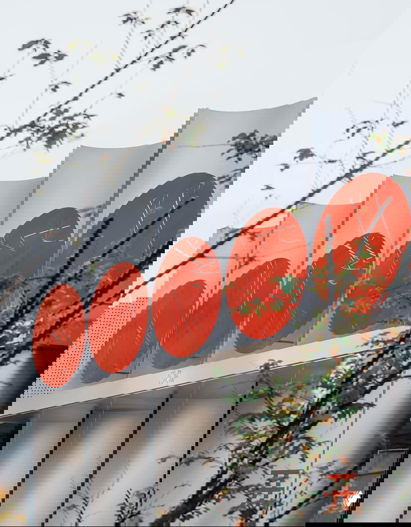 Фрагмент фасада в ракурсе: крупные красные диски на серебристых полуцилиндрах, ветви деревьев на переднем плане подчёркивают связь с парком.