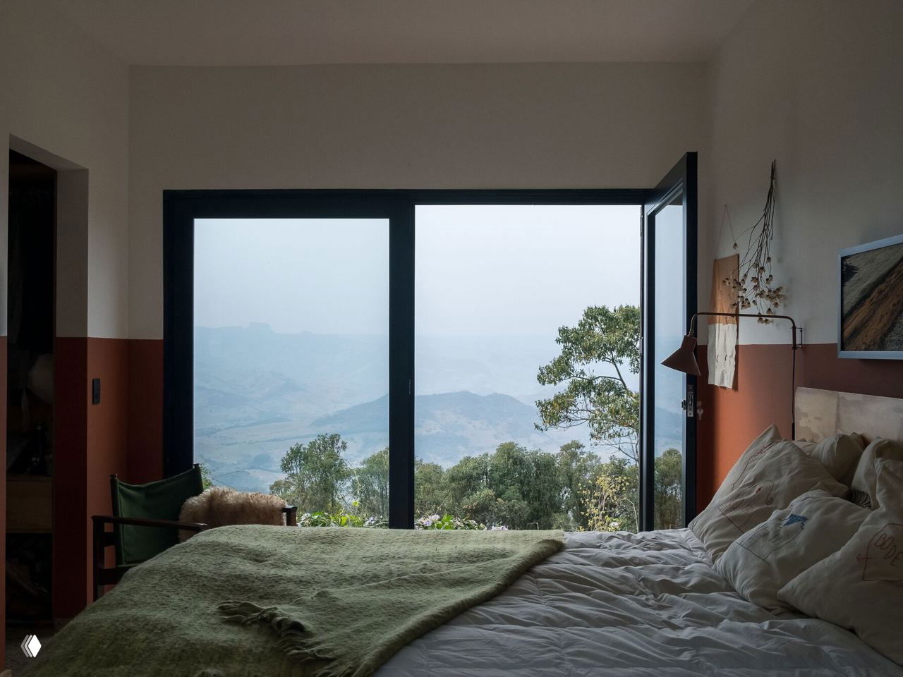 Уютная спальня с раздвижным панорамным остеклением и видом на холмы; натуральные текстуры и приглушённые цвета подчёркивают связь с природой за окном.
