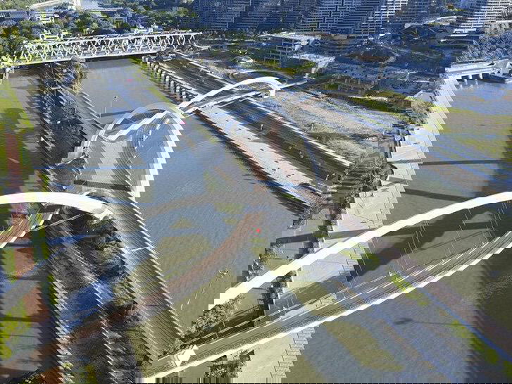 План сверху: три переплетённые арки пешеходно‑велосипедного моста над узким участком канала, видны насыпной остров и дорожные переходы.