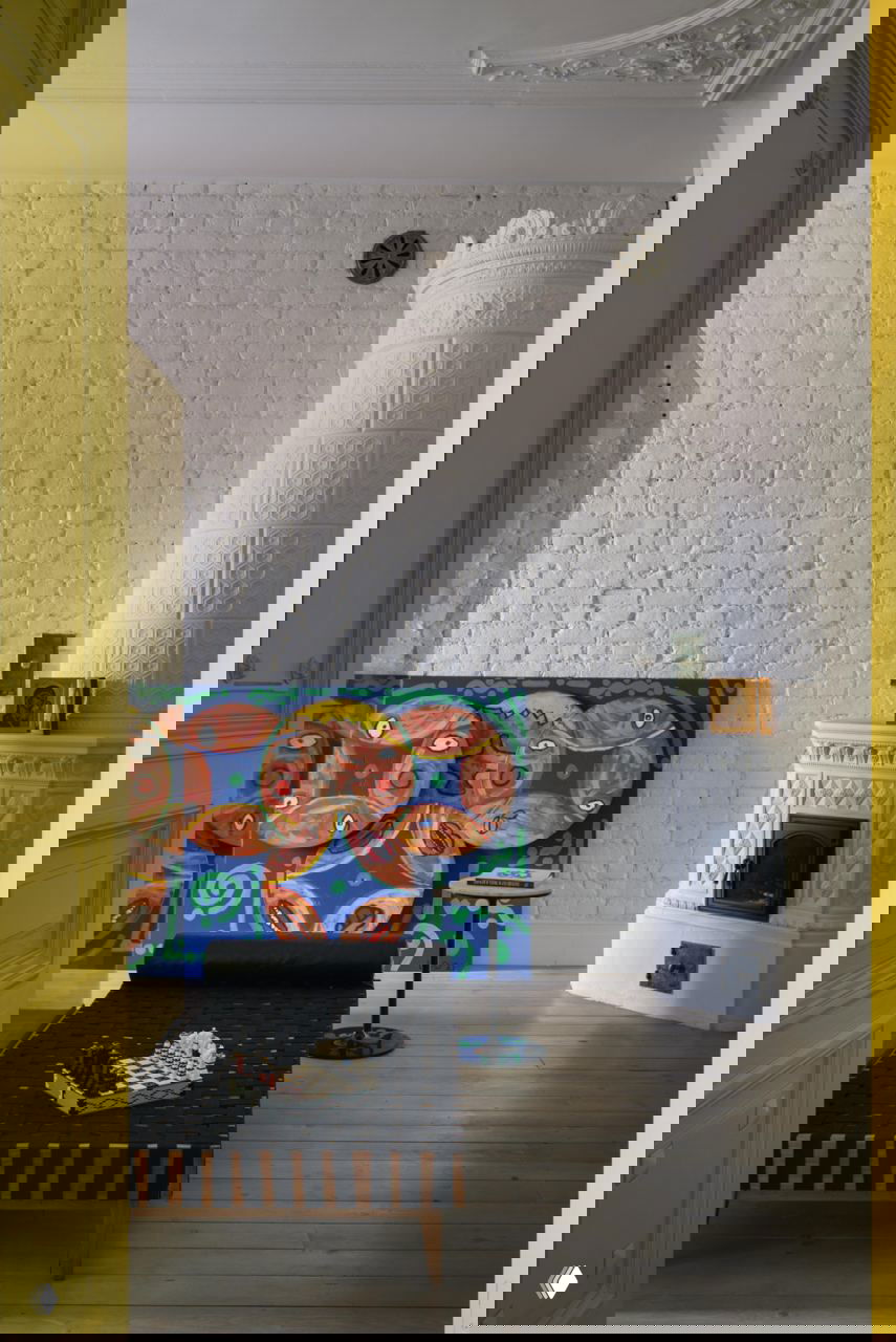 Городской интерьер с белой расписной изразцовой печью на фоне кирпичной стены и яркой живописной картины, контрастные жёлтые дверные откосы в кадре.