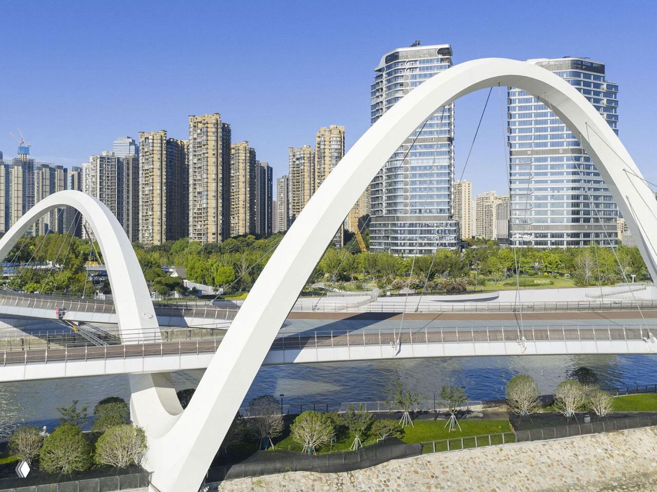Крупный план белых стальных арок моста на фоне жилых высоток Ханчжоу: конструкция и опоры набережной в дневном свете.