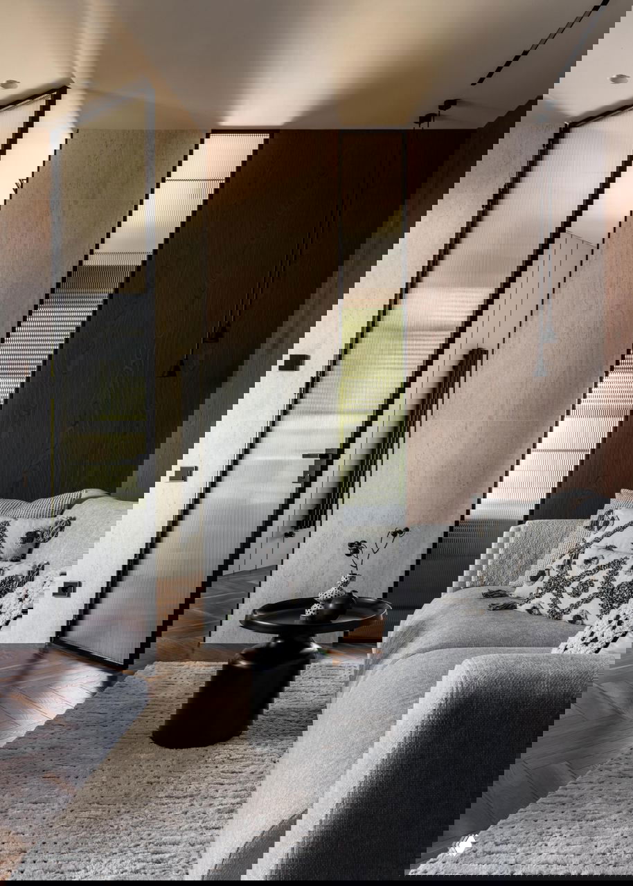 Вид на зону гостиной: светлый диван, декоративные подушки, низкий чёрный столик и узкие вертикальные окна с рифлёным стеклом на фоне деревянной стеновой панели.